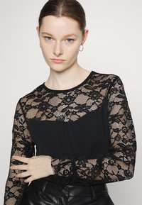 Top preto com mangas longas em renda preta translúcida com padrões florais. O corpo é de um tecido sólido preto com decote redondo e mangas longas.