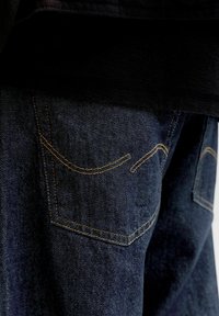 Rückansicht einer Person, die dunkelblaue Jeans mit gelber Naht an den Gesäßtaschen trägt, darüber teilweise sichtbar ein schwarzes Hemd.