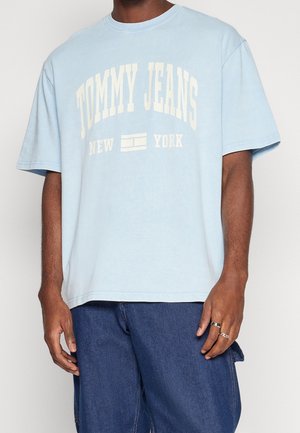 Osoba ubrana w jasnoniebieską koszulkę z napisem i logo "TOMMY JEANS NEW YORK", w połączeniu z ciemnoniebieskimi dżinsami i srebrnymi pierścieniami na prawej ręce.