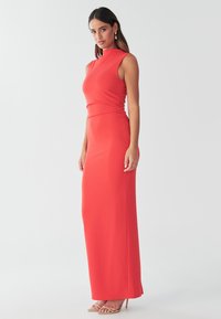 Lang, figursyet rød kjole lavet af elastisk stof med højt halsudskæring og rynkede detaljer i siden, kombineret med nøgenfarvede, snørede hæle.