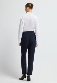 Bílá košile s dlouhým rukávem a límečkem, zkombinovaná s tmavě modrými upnutými kalhotami a černými texturovanými vysokými podpatky, viděná zepředu.