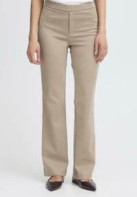 Pantaloni beige a zampa d'elefante realizzati in tessuto morbido con una texture liscia. Presentano una parte anteriore piatta e un design sobrio, abbinati a scarpe marroni con punta.