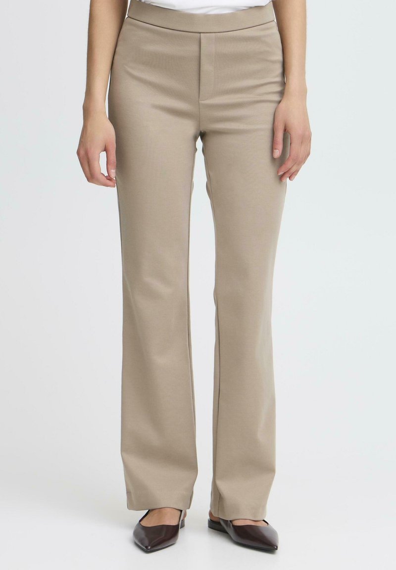 Pantaloni beige a zampa d'elefante realizzati in tessuto morbido con una texture liscia. Presentano una parte anteriore piatta e un design sobrio, abbinati a scarpe marroni con punta.