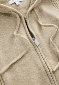 Beige vetoketjullinen huppari, joka on valmistettu tekstuurilla kudotusta kankaasta. Se sisältää hopeanvärisen vetoketjun ja nyörit, sekä ribbireunat kaula-aukolla.