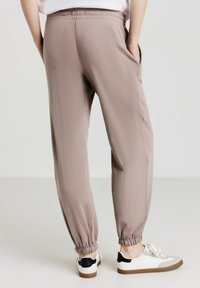 Helle taupe Jogginghosen aus weichem Stoff mit elastischem Bund und Bündchen, ausgestattet mit Seitentaschen und lockerer Passform. Darunter sind legere Schuhe.