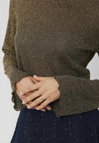 Haut à manches longues pailleté marron avec une finition texturée et des poignets festonnés, porté avec une jupe à motif foncé. Mains jointes à la taille.