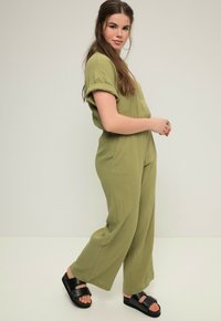 Olivengrüner, leger geschnittener Jumpsuit mit kurzen Ärmeln, Knopfverschluss, seitlichen Taschen und strukturiertem Material. Kombiniert mit schwarzen Plateausandalen.