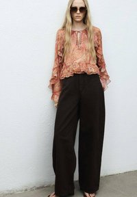 Bloemige geplooide blouse in oranje en roze tinten, gecombineerd met wijde, donkerbruine broeken. Accessoires zijn ronde zonnebrillen en sandalen.