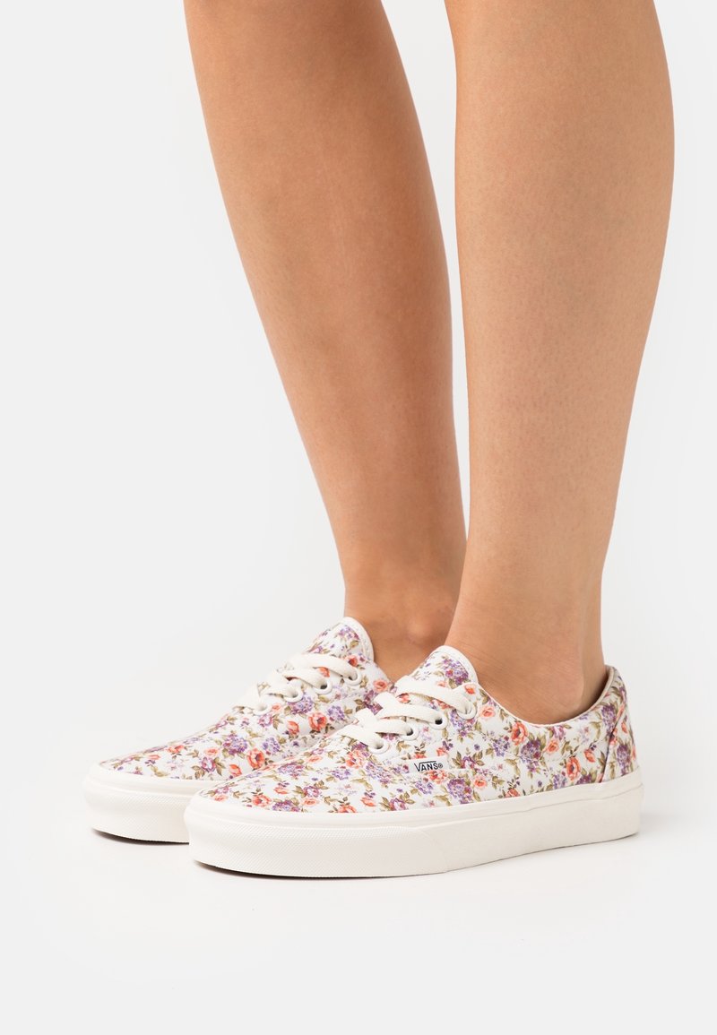 Deux pieds portant des baskets Vans blanches à lacets avec un motif floral multicolore sur fond blanc.