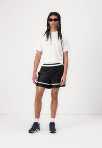 Shorts de sport noirs avec des bandes blanches sur les côtés, associés à un t-shirt blanc uni. Portés avec des baskets sombres et des lunettes de soleil noires.