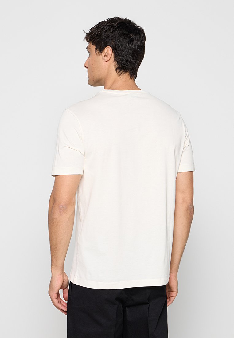 Hombre con cabello corto y oscuro, vestido con una camiseta blanca lisa de manga corta y pantalones negros, de espaldas sobre un fondo gris claro.