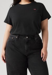 Woman wearing a plain black Levi's t-shirt tucked into high-waisted black denim jeans, standing against a light gray background.