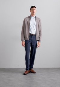 Cárdigan marrón claro sobre una camisa de botones azul claro, combinado con pantalones azul oscuro y zapatos marrones, todo sobre un fondo sencillo.