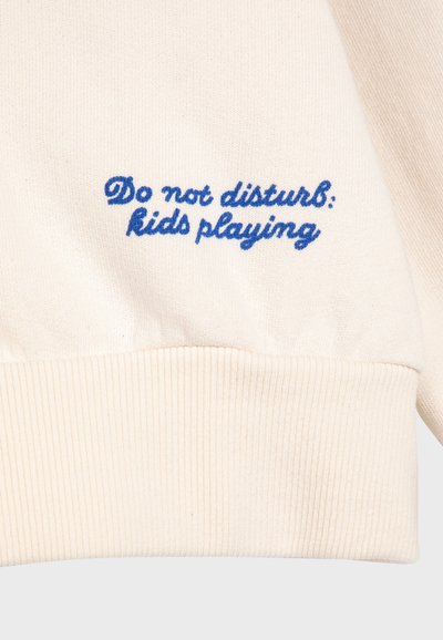 Krémový pletený sveter s modrým vyšívaným nápisom "Nerušiť: Hrajúce si deti" pri manžete. Má rebrovaný lem.