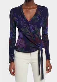 Mujer vestida con una blusa envolvente de flores en tonos púrpuras oscuros y mangas largas, con pantalones color beige claro, la blusa atada en la cintura por el lado derecho.