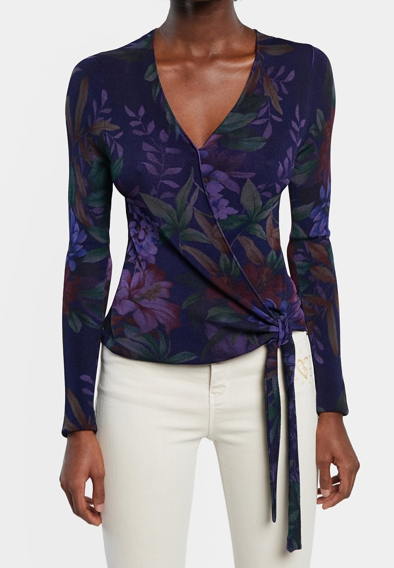 Mujer vestida con una blusa envolvente de flores en tonos púrpuras oscuros y mangas largas, con pantalones color beige claro, la blusa atada en la cintura por el lado derecho.