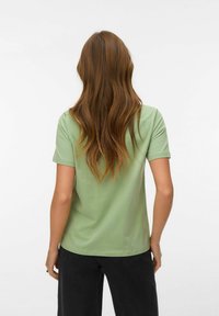 Γυναίκα με μακριά κυματιστά καστανά μαλλιά, φοράει ανοιχτό πράσινο κοντομάνικο πουκάμισο και μαύρο παντελόνι, στέκεται αντικρίζοντας ένα απλό λευκό φόντο.