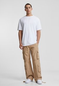 Tricou alb cu guler rotund, purtat cu pantaloni cargo bej care au multiple buzunare laterale și adidași albi. Design minimalist, croială lejeră.