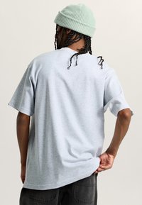 Světle šedé oversized tričko vyrobené z měkkého materiálu, s krátkými rukávy a kulatým výstřihem. Nosené s tmavě šedými kalhotami a světle zeleným čepichem.