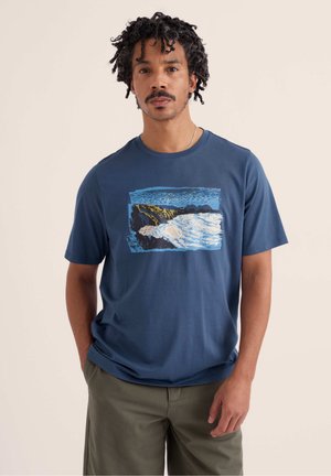 Mann mit lockigem Haar trägt ein blaues T-Shirt mit Motiv von Ozeanwellen, die gegen felsige Klippen schlagen, kombiniert mit olivgrünen Hosen.