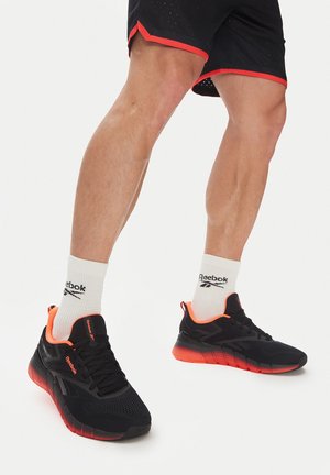 Zapatillas deportivas Reebok negras con parte superior de malla, acentos naranjas y suela texturizada, llevadas con calcetines blancos que presentan el logo de la marca.