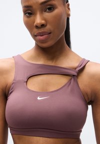 Reggiseno sportivo viola in tessuto liscio con design traforato. Presenta una sottile fascia sul petto e un logo Nike bianco sul davanti.
