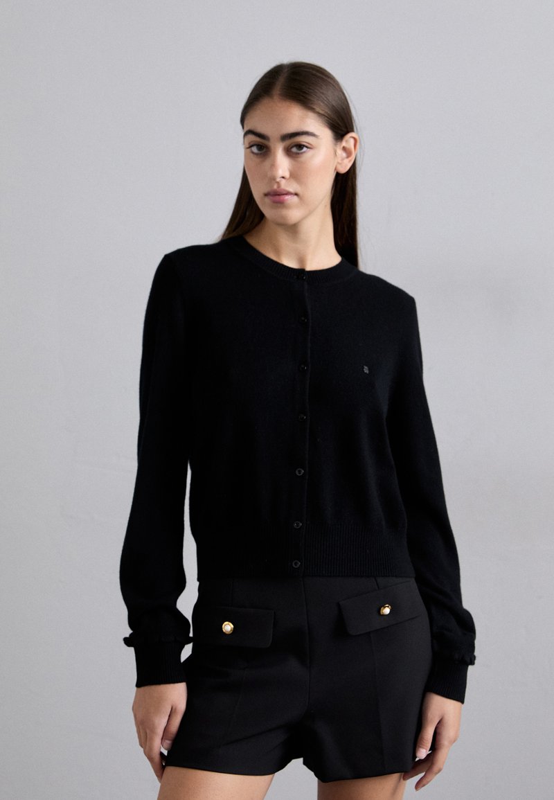Cardigan in lana nera con scollatura rotonda, bottoni frontali e polsini a costine, abbinato a pantaloni corti neri a vita alta con dettagli dorati.