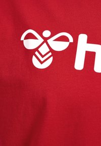 Rød bomuldsstof med et stort hvidt bi-logo med stiliserede vinger og geometriske mønstre, sammen med hvid tekst.