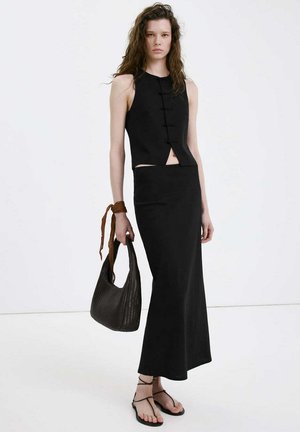 Kobieta w czarnym topie bez rękawów i długiej czarnej spódnicy, trzyma ciemną plecioną torebkę z brązową apaszką zawiązaną wokół nadgarstka.