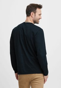 Mustas pikkade varrukatega särk ümmarguse kaelusega, relaxed fit ja tekstureeritud kangaga. Omab väikest tagumiste sildiga detaili ja on kombineeritud beežide pükstega.