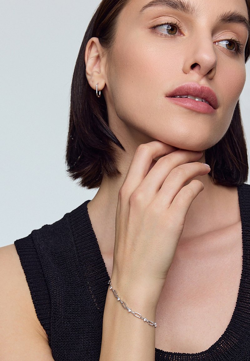 Srebrna bransoletka z wydłużonymi i kanciastymi ogniwami, ozdobiona małymi akcentami kryształowymi. Noszona na nadgarstku, w zestawieniu z ciemnym swetrem.