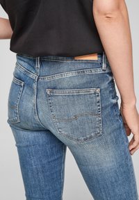 Person wearing light blue denim jeans and a black shirt, shown from the waist down, facing away with one arm by their side.