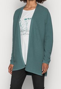 Mujer vestida con un cárdigan abierto de canalé en color verde azulado sobre una camiseta gráfica blanca con flores y pantalones negros, de pie con las manos relajadas a los lados.
