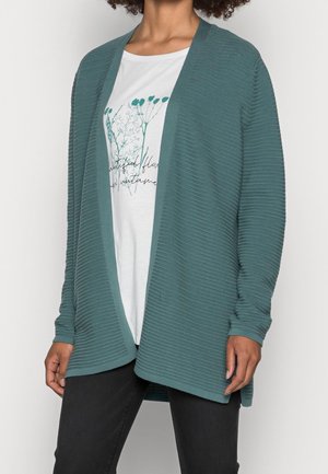 Femme portant un cardigan ouvert en ribbed teal sur un tee-shirt graphique blanc à fleurs et un pantalon noir, se tenant avec les mains détendues le long du corps.
