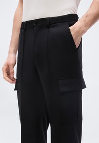 Άνδρας που φοράει μαύρο παντελόνι cargo με πλαϊνές τσέπες με καπάκια και το ένα χέρι στην τσέπη, σε απλό ανοιχτό φόντο.