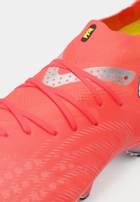 Jasnoróżowy but piłkarski z teksturowaną powierzchnią, srebrnymi akcentami i czerwonymi sznurowadłami. Charakteryzuje się smukłym designem z niskim profilem i podeszwą zapewniającą przyczepność.