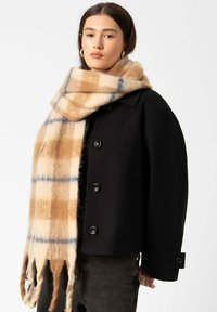 Zwarte wollen jas met grote knopen, vergezeld van een sjaal met een ruitpatroon in beige, bruin en blauwe tinten. Lange franjes aan de sjaal.