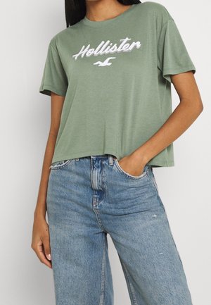 Person iført en løs grøn Hollister t-shirt med korte ærmer, let stoppet ind i høj talje lyseblå jeans.