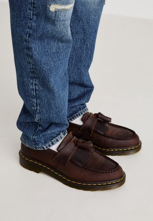 Persoon draagt blauwe spijkerbroek en bruine leren loafers met franjes, gele stiksels en witte sokken op een lichte vloer.