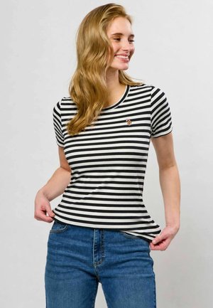 Femme aux longs cheveux blonds sourit en tenant l'ourlet de son t-shirt à manches courtes rayé noir et blanc, portant un jean bleu.
