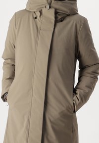 Beige, isoleret lang frakke med høj krave, hætte og lynlås. Har sidelommer og ribstrikkede manchetter, lavet af et glat materiale.