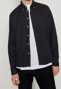 Camisa de botones en chambray oscuro con botones dorados, mangas largas y un cuello clásico. Usada sobre una camiseta blanca de cuello redondo.