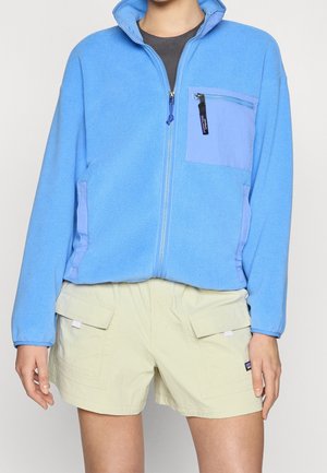 Person trägt hellblaue Fleecejacke mit Frontreißverschluss und Reißverschlusstasche, kombiniert mit beigen Cargoshorts mit Klappentaschen.