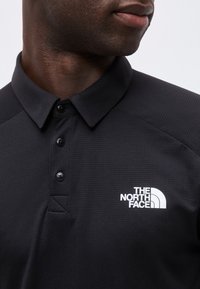 Gros plan d'une personne portant un polo noir à col avec le logo "The North Face" en blanc sur le côté gauche de la poitrine.