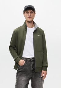 Zöld, cipzáras kapucnis pulóver magas gallérral, szürke béléssel és hímzett logóval. Fehér ing fölött viselve, szürke farmernadrággal párosítva.