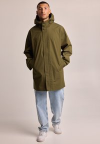 Olive green waterproof jacket with a hood, simple button closure, and pockets, paired with light blue jeans and white sneakers.