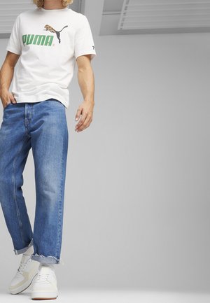 Vit t-shirt med korta ärmar, med en grön "PUMA"-logotyp och leopardgrafik; kombinerad med ljusblå jeans och vita sneakers.