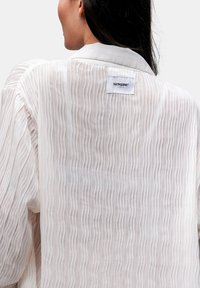 Blusa leggera plissettata in morbido beige con colletto bianco. Presenta un'etichetta cucita sul retro con il nome del marchio. Il tessuto testurizzato crea profondità.