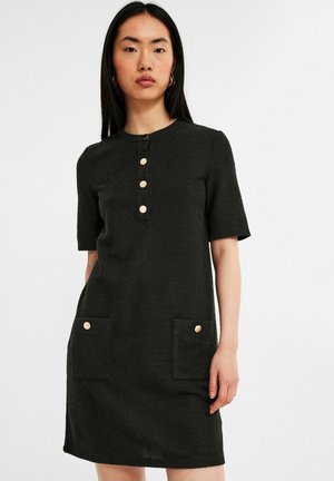 Woman with long black hair wearing a short-sleeve black textured dress with gold buttons and front pockets against a plain white background.