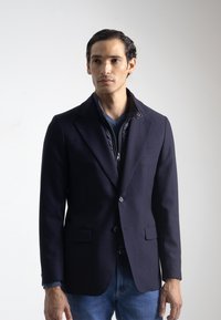 Chaqueta náutica con tejido texturizado, solapas con muesca, dos bolsillos frontales y un cuello acolchado en capas, llevada sobre una camiseta azul de cuello en V.